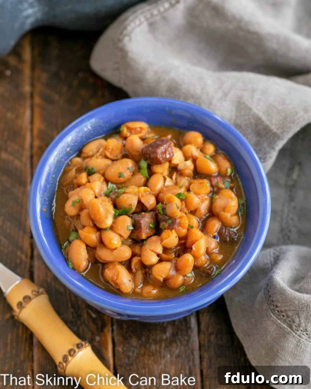 Grandma's Classic Baked Beans 3 homemade baked beans in a blue bowl with a bamboo fork to the lower left.