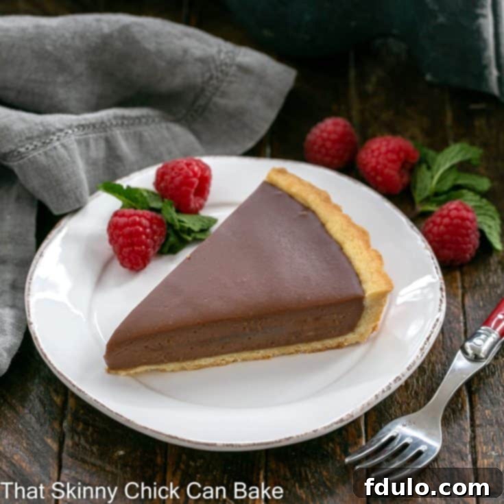 A wedge of milk chocolate tart on a white dessert plate with raspberry garnish and a red handle fork