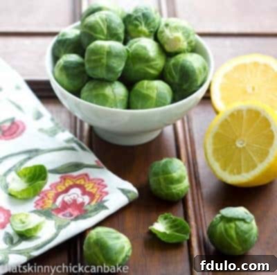 Fresh Brussels Sprouts in a small white bowl positioned next to neatly sliced lemons on a rustic wooden table, illustrating fresh ingredients.