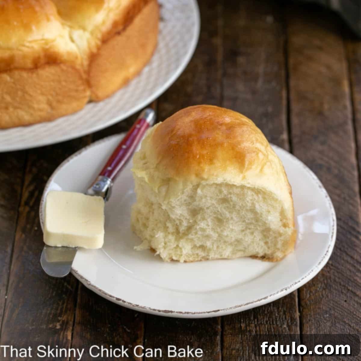 Soft and Fluffy Japanese Milk Bread 4 A single, perfectly golden-brown Japanese Milk Bread Roll on a white plate, accompanied by a red handled knife and a melting pat of butter, inviting a delicious bite.