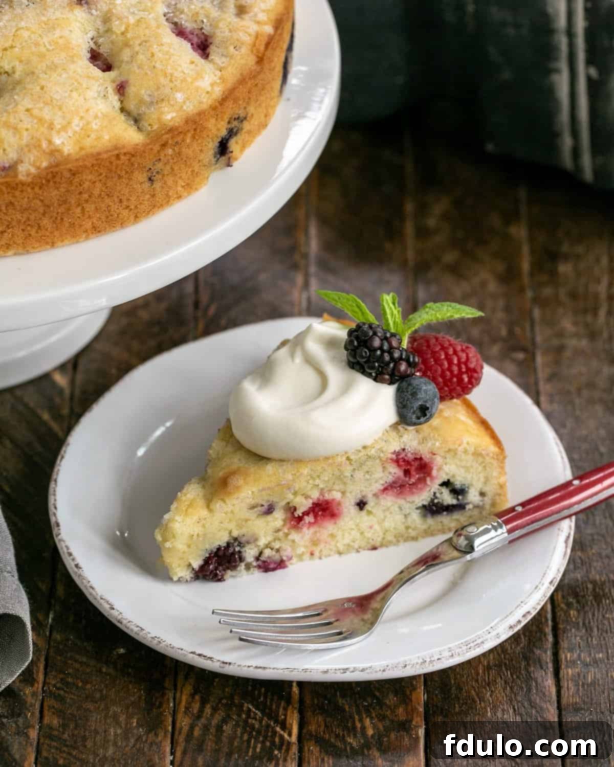A perfectly sliced berry cake on a white plate, topped with a generous dollop of lemon curd cream and a few fresh berries, ready to be enjoyed with a red-handled fork.