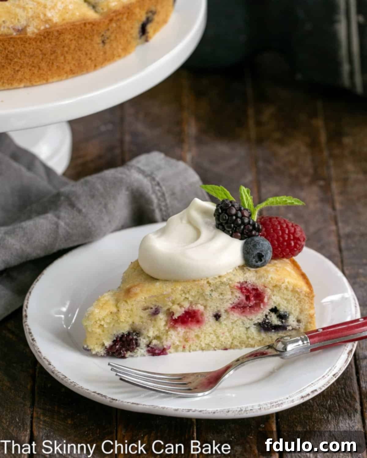 A slice of berry cake topped with fluffy lemon whipped cream and fresh berries, placed on a white plate in front of a full cake on a stand.