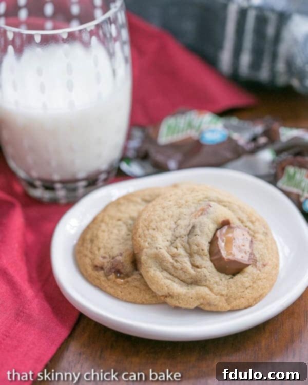 Two warm Milky Way Cookies with visible chunks of Milky Way candy on a small white plate, accompanied by a refreshing glass of milk.