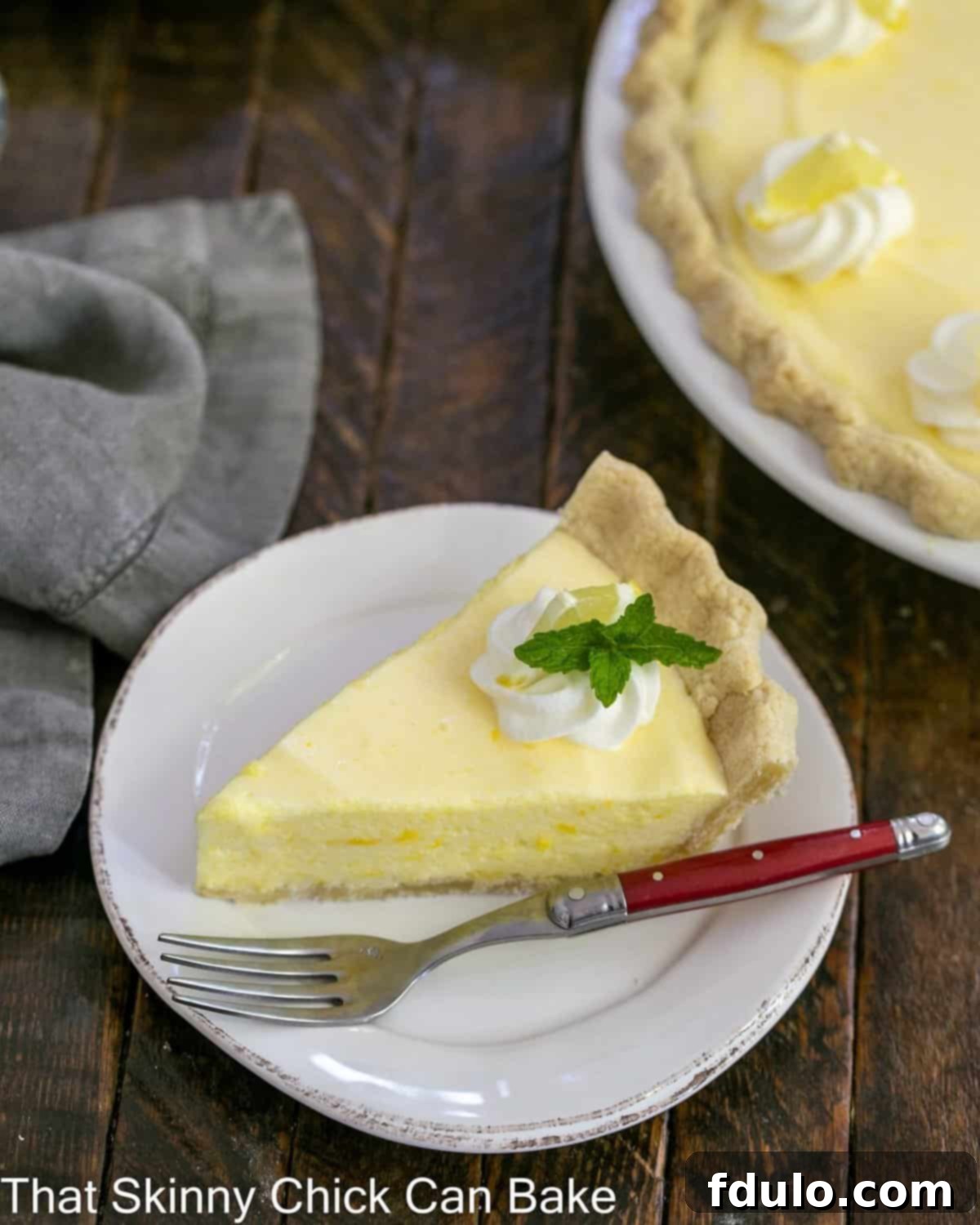 Slice of lemon chiffon pie on a white plate with a red handle fork and whole pie in the background.