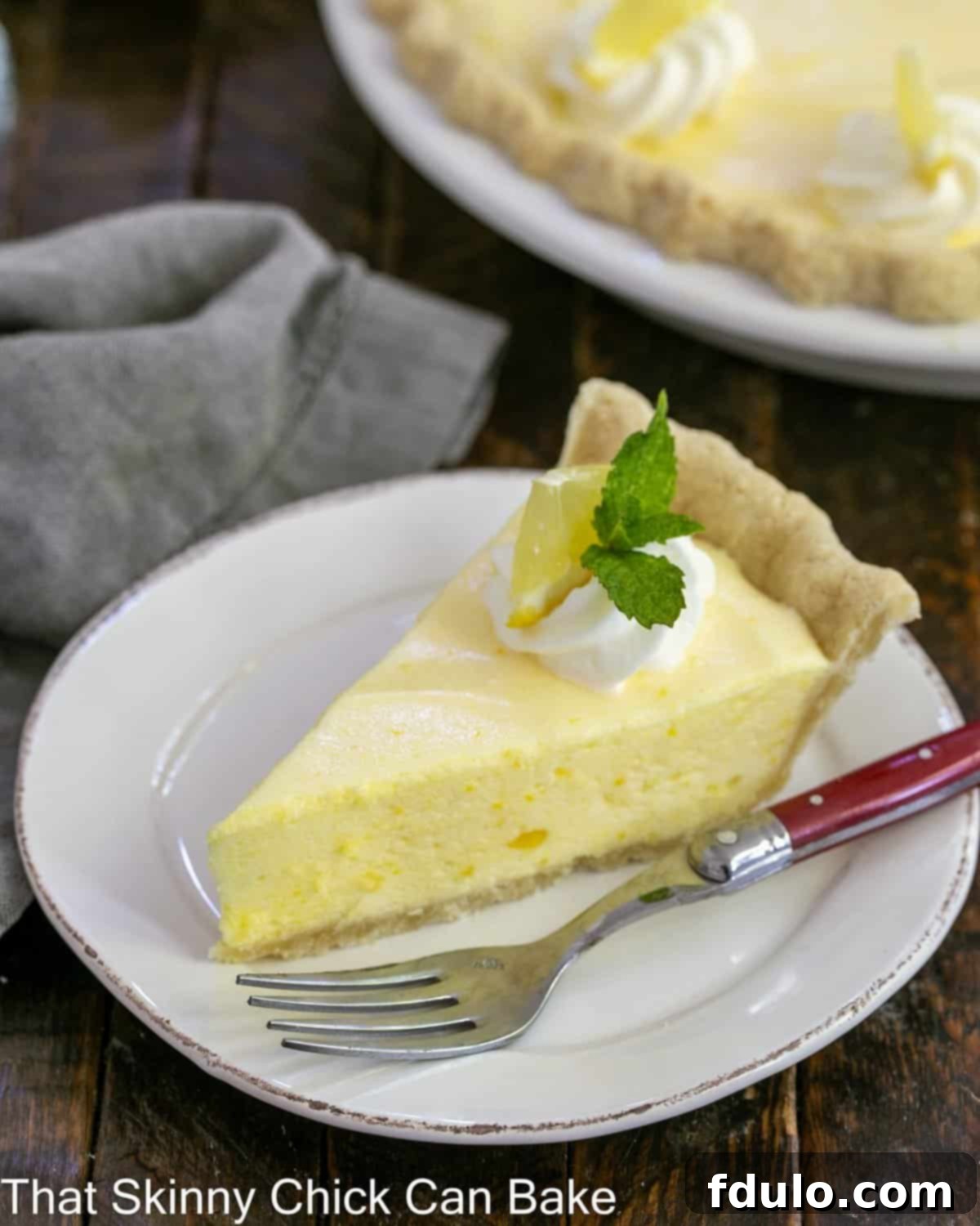 Slice of lemon chiffon pie on a white dessert plate with a swirl of whipped cream and mint sprig garnish.