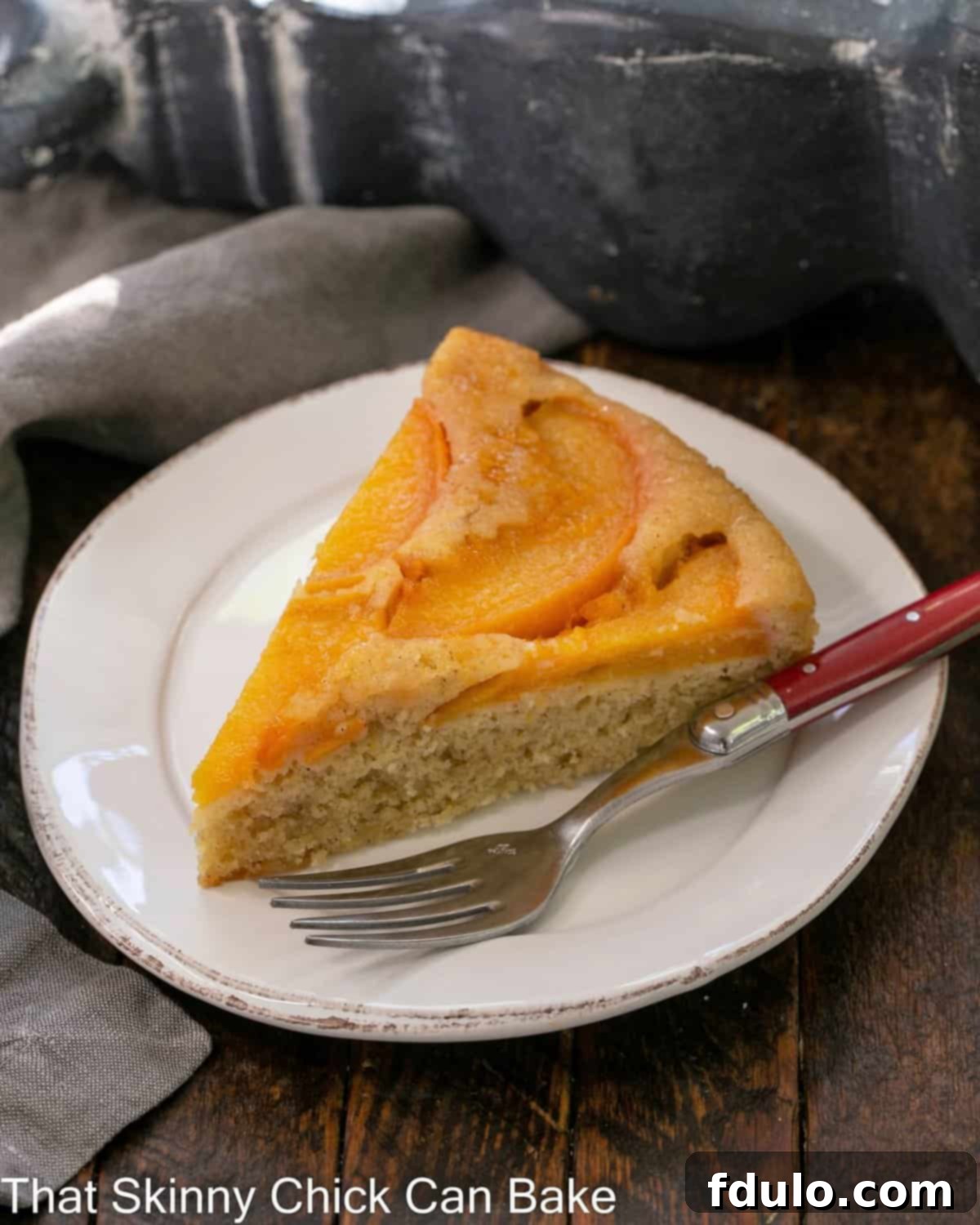 A slice of peach upside down cake on a white dessert plate with a red handle fork, showcasing the caramelized peach topping and moist cake.