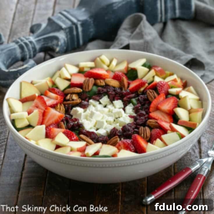 Spinach salad with strawberries and feta in a white ceramic salad bowl and 2 serving utensils.