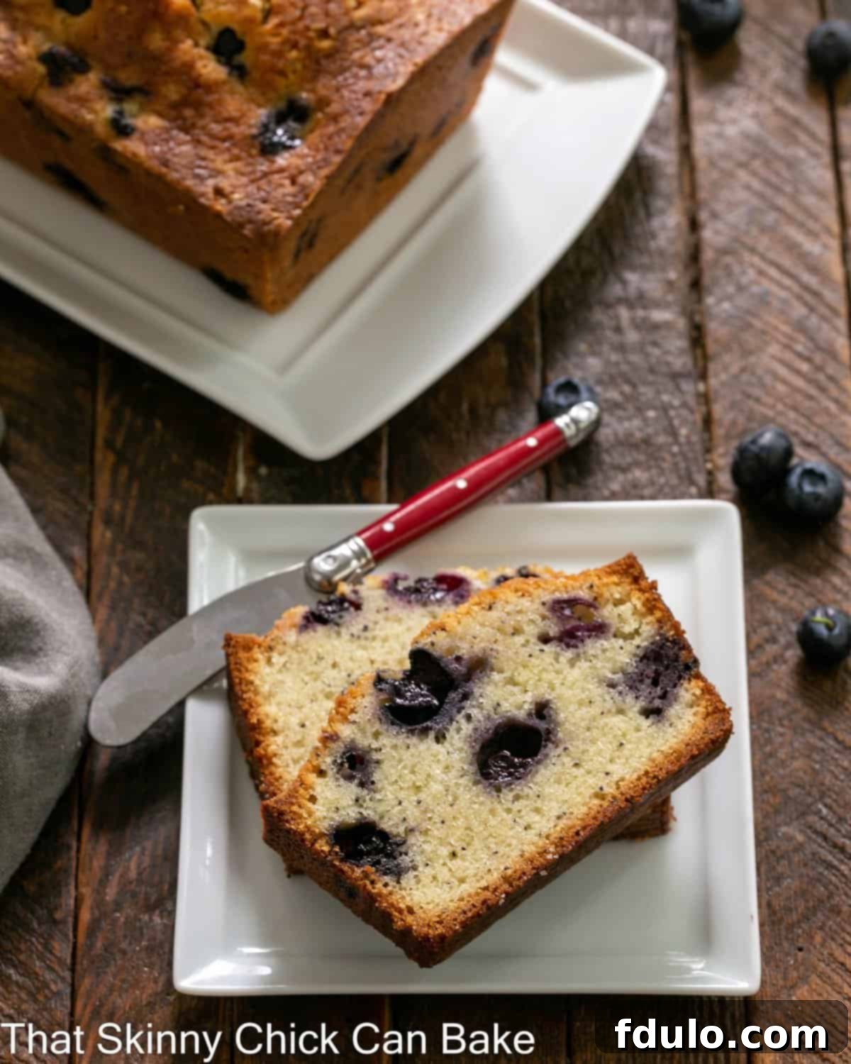Two slices of blueberry bread on a white plate in front of a loaf on a serving tray.