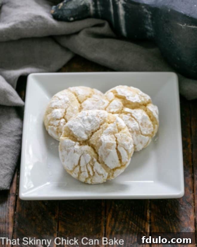 Three golden-brown Gooey Butter Cookies with a delicate crackled surface, dusted with powdered sugar, resting on a square white plate, hinting at their soft, chewy interior.