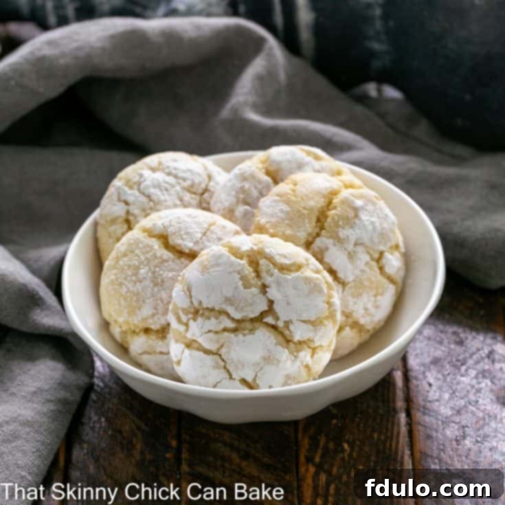 A small white ceramic bowl filled with five soft, golden Gooey Butter Cookies, with a sixth cookie peeking out from behind, all dusted with a generous layer of powdered sugar.
