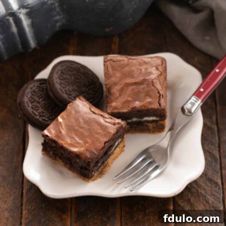 Chocolate Chip Oreo Brownies 14 Two decadent Chocolate Chip Oreo Cookie Brownies served on a pristine white plate, accompanied by a fork with a red handle, inviting you to take a bite.