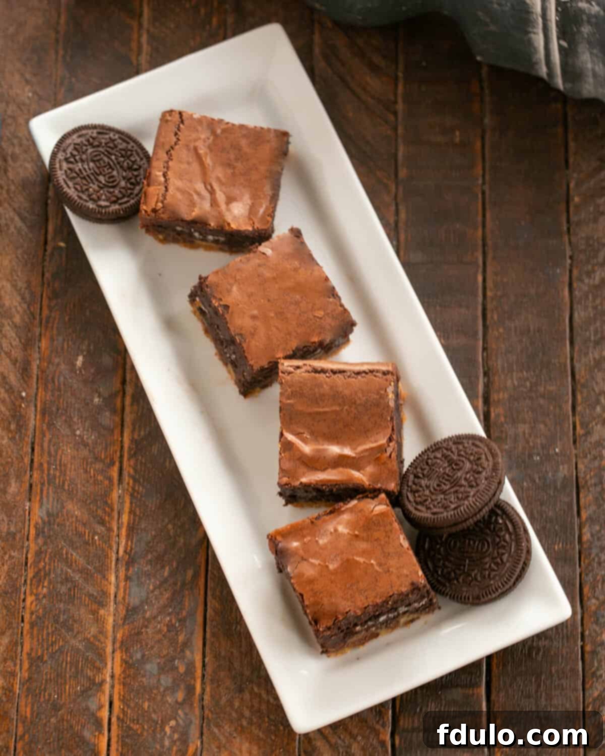 Chocolate Chip Oreo Brownies 3 Four perfectly portioned Chocolate Chip Oreo Cookie Brownies arranged on a elegant white ceramic tray, complemented by several whole Oreo cookies and halves, ready to be served and enjoyed.