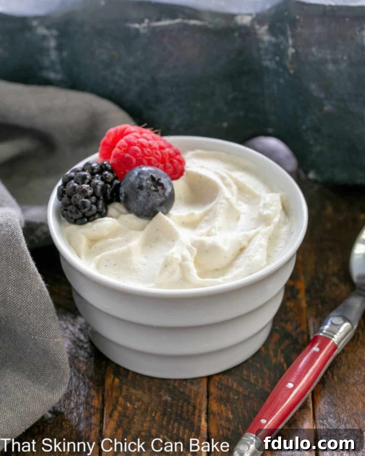 Crème Chantilly in a white ramekin topped with 3 berries next to a red handled spoon.