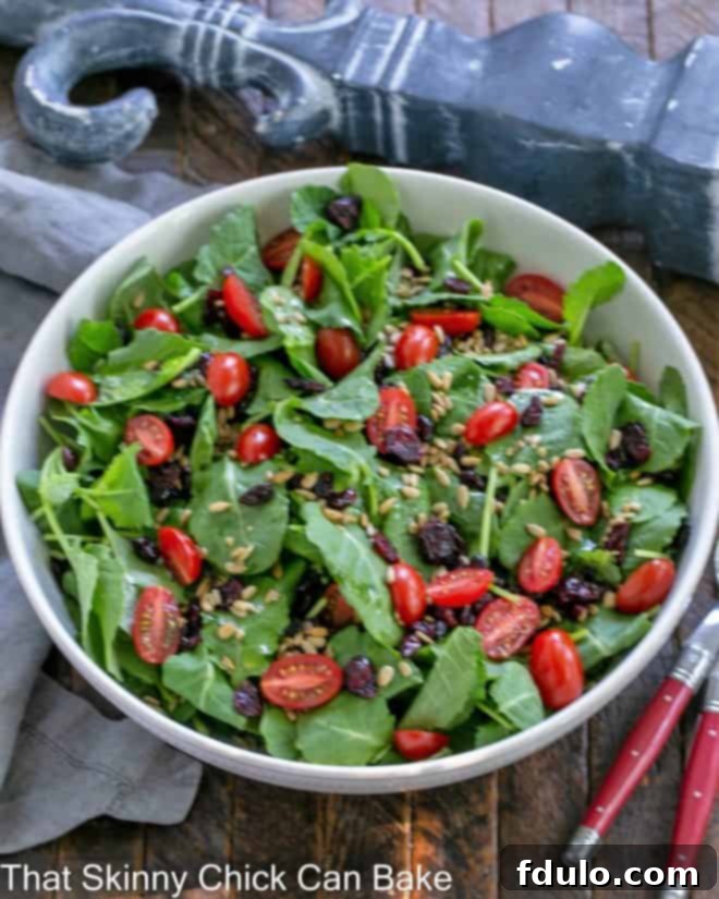 An inviting overhead shot of a fresh kale salad, brimming with cranberries, tomatoes, and sunflower seeds, served in a pristine white bowl with elegant red-handled serving utensils.