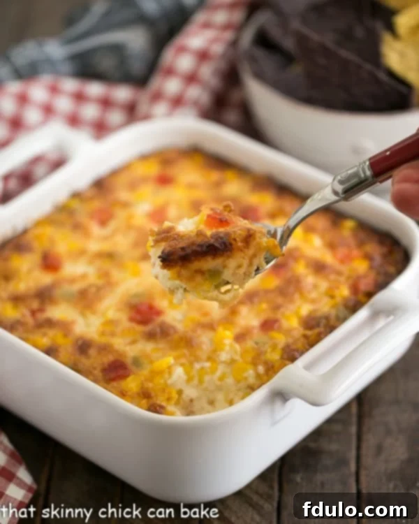 Fiesta Corn Dip - With a double dose of gooey cheese, you'll want to serve this magnificent dip at every gathering! A serving spoon lifting a generous scoop of hot, gooey Fiesta Corn Dip from a white casserole dish, showcasing its cheesy texture.