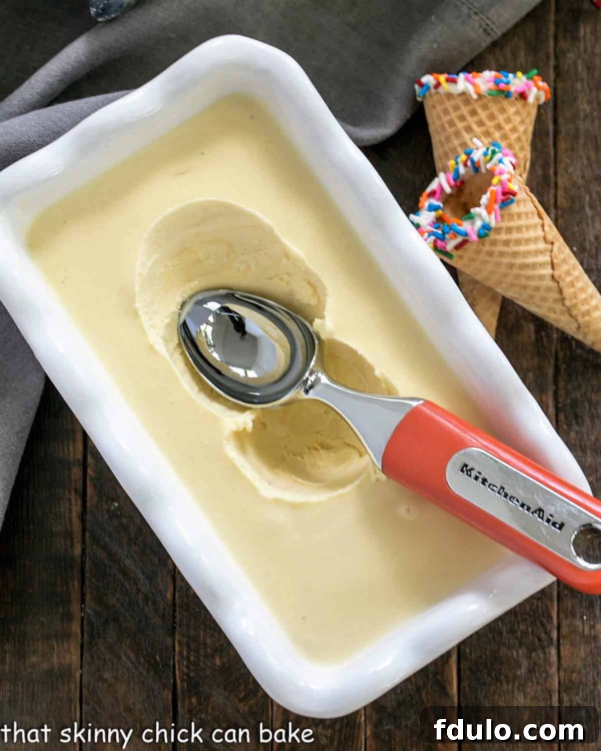 A large tub of freshly churned homemade cake batter ice cream in a freezer-safe dish, with a metal ice cream scooper resting on the edge, ready for serving.