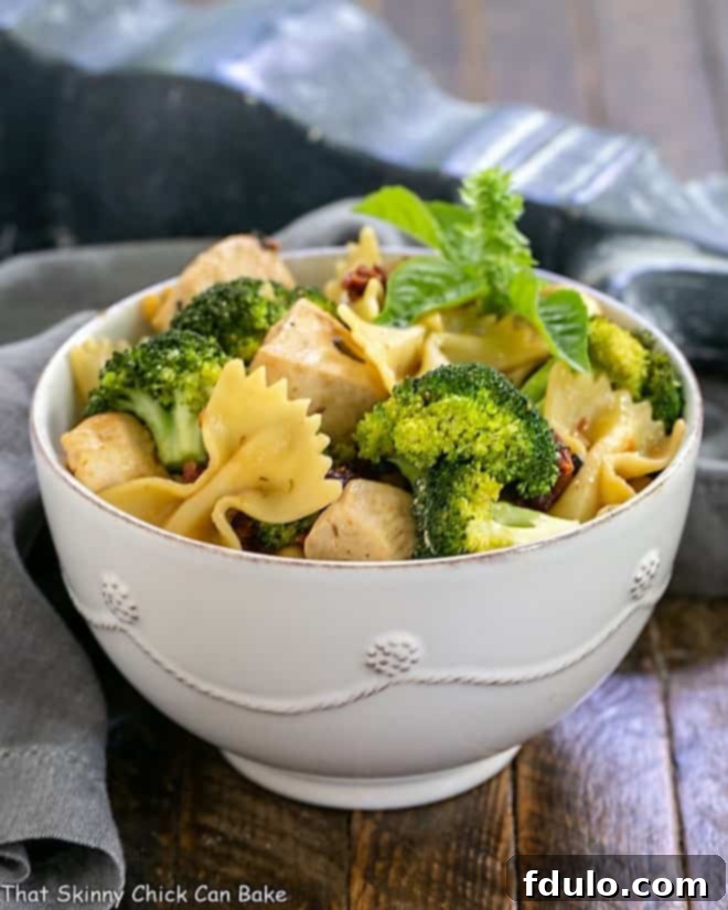 A close-up shot of a white ceramic bowl filled with Chicken with Broccoli, Sun-dried Tomatoes and Bow Tie Pasta, garnished with fresh herbs, showcasing the vibrant colors and appealing texture of the dish.