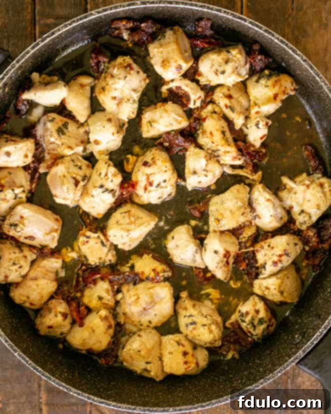 Overhead view of cubed chicken sautéing in a skillet with minced garlic, dried basil, red pepper flakes, seasoning salt, and vibrant sun-dried tomato strips.