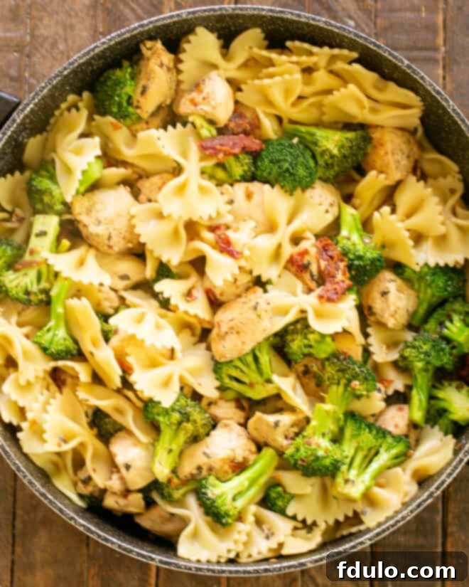 Cooked bow tie pasta being added to the skillet with the chicken, broccoli, and sun-dried tomato sauce, ready to be tossed and coated in the delicious juices.