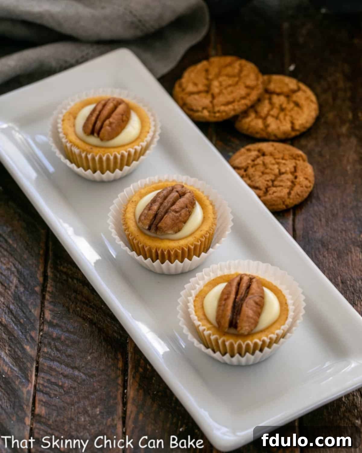 Spiced Pumpkin Cheesecake Cups 2 Three perfectly formed mini pumpkin cheesecakes arranged on a crisp white tray, with a scattering of ginger cookies in the background, hinting at their delicious crust.