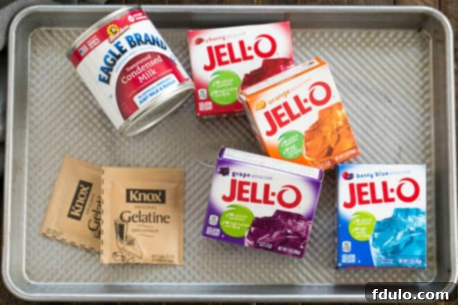 Stained Glass Dessert 3 Ingredients for Broken Glass Jello laid out on a sheet pan, including various colored Jello boxes, unflavored gelatin, and sweetened condensed milk.