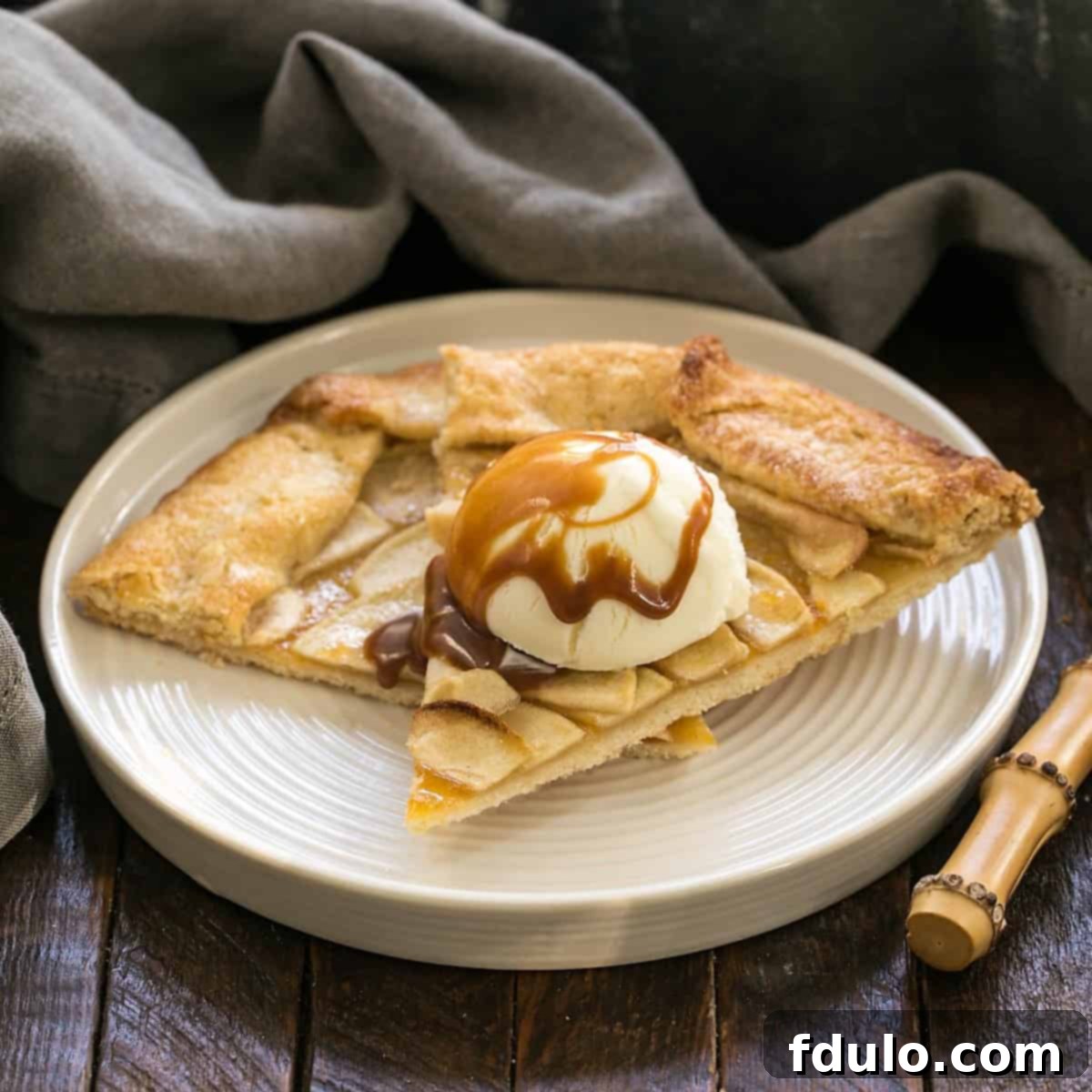 Two generous slices of homemade Apple Crostata, served with a scoop of vanilla ice cream and a drizzle of rich caramel sauce, on a rustic wooden surface.