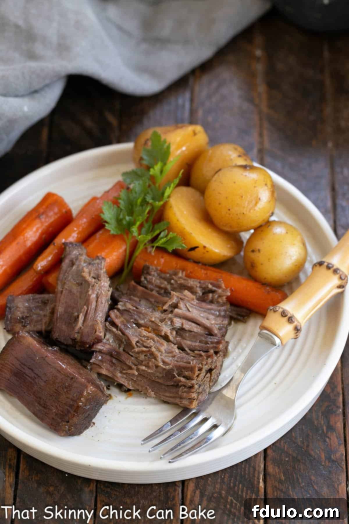Tender Beef Chuck Roast 3 Beef pot roast on a white dinner plate with carrots, potatoes, and a bamboo handle fork, garnished with fresh herbs.