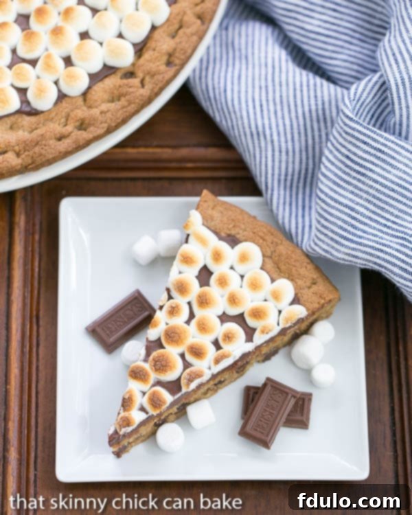 A delectable wedge of S'mores Cookie Cake presented on a clean, square white dessert plate, highlighting its distinct layers of cookie, chocolate ganache, and toasted marshmallows.