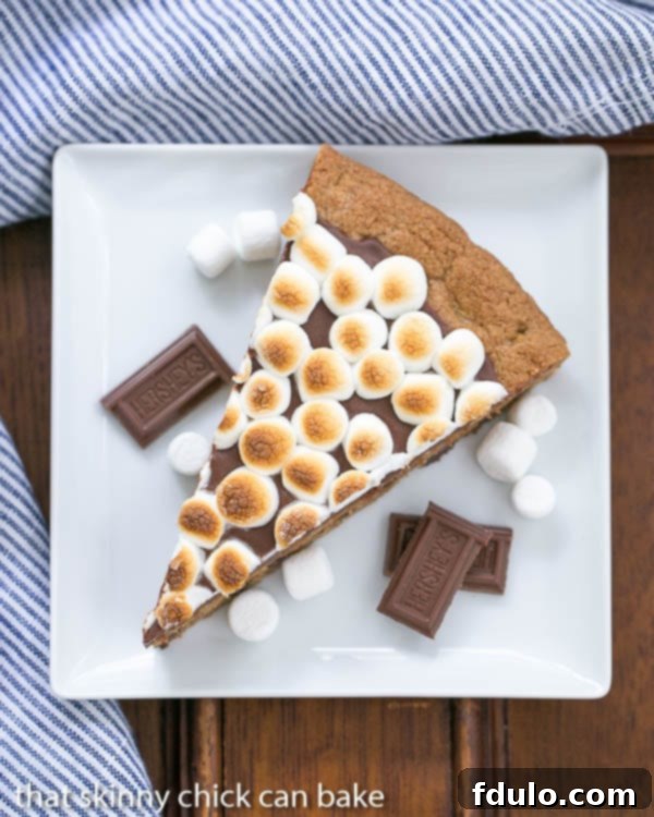 A perfectly sliced wedge of S'mores Cookie Cake on a white plate, garnished with extra chocolate bar squares and mini marshmallows for an inviting presentation.