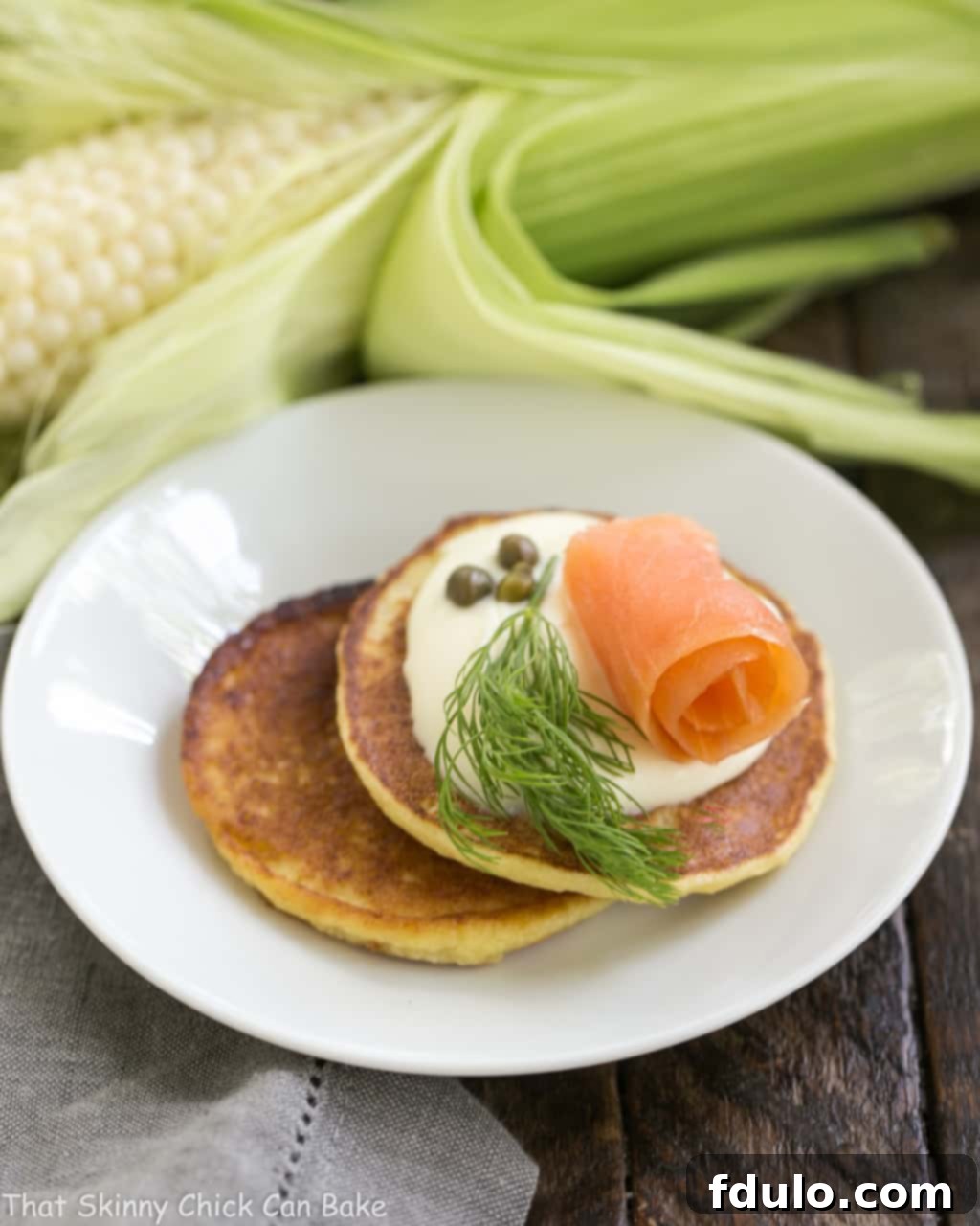 Savory Corn Cakes beautifully arranged on a round white plate with fresh herbs and a dollop of creme fraiche.