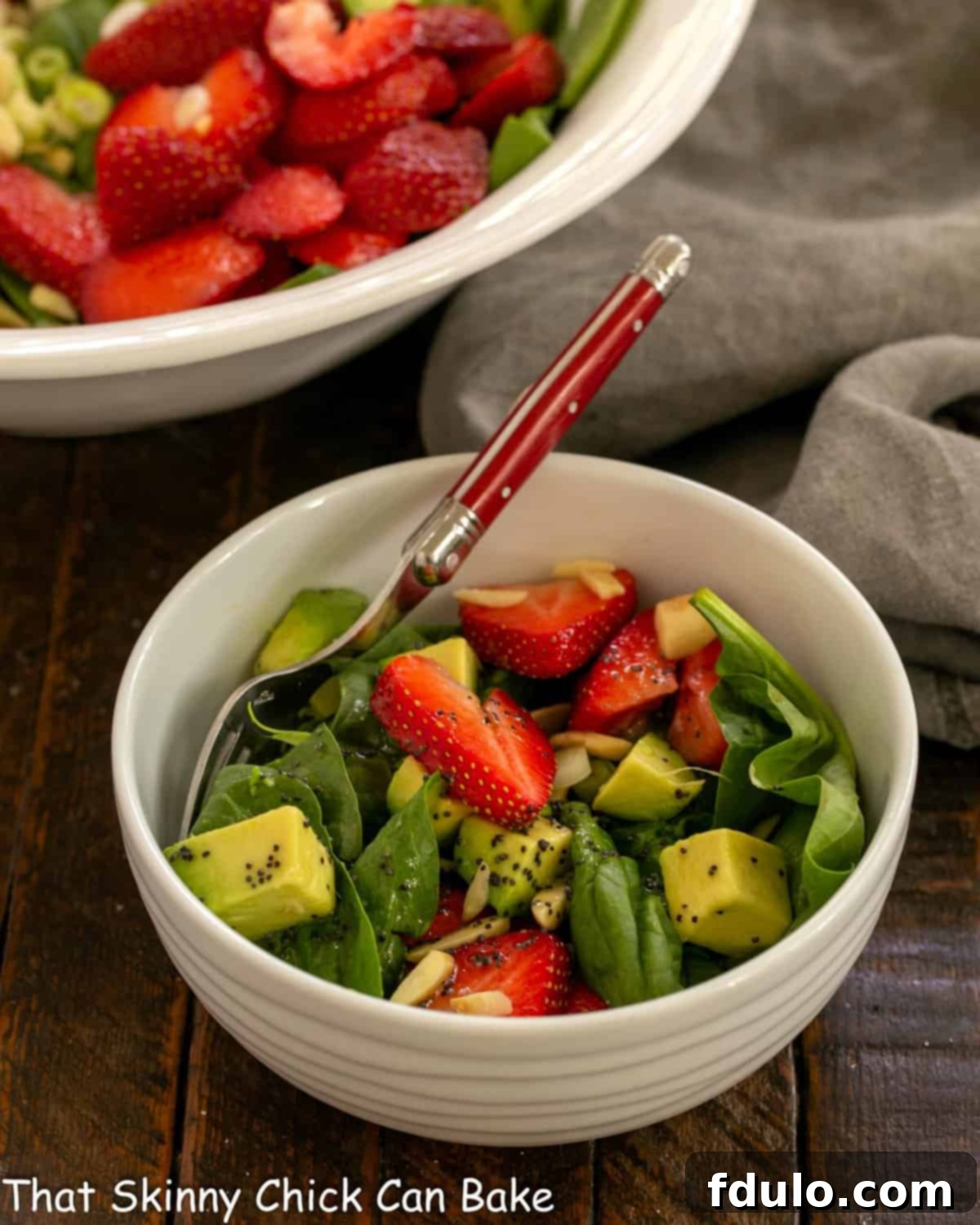 A vibrant Strawberry Spinach Salad with Avocados served in a small white ceramic bowl, ready to be enjoyed with a red-handled fork.
