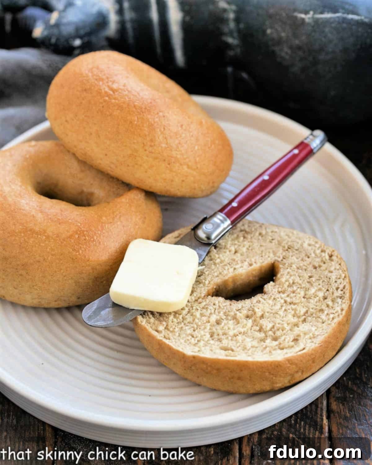 Whole Wheat Bagels - Dense, chewy and delicious and made in your own kitchen! Two golden-brown whole wheat bagels resting on a pristine white plate, with one sliced open to reveal its tender interior, accompanied by a small pat of butter and a red-handled knife.