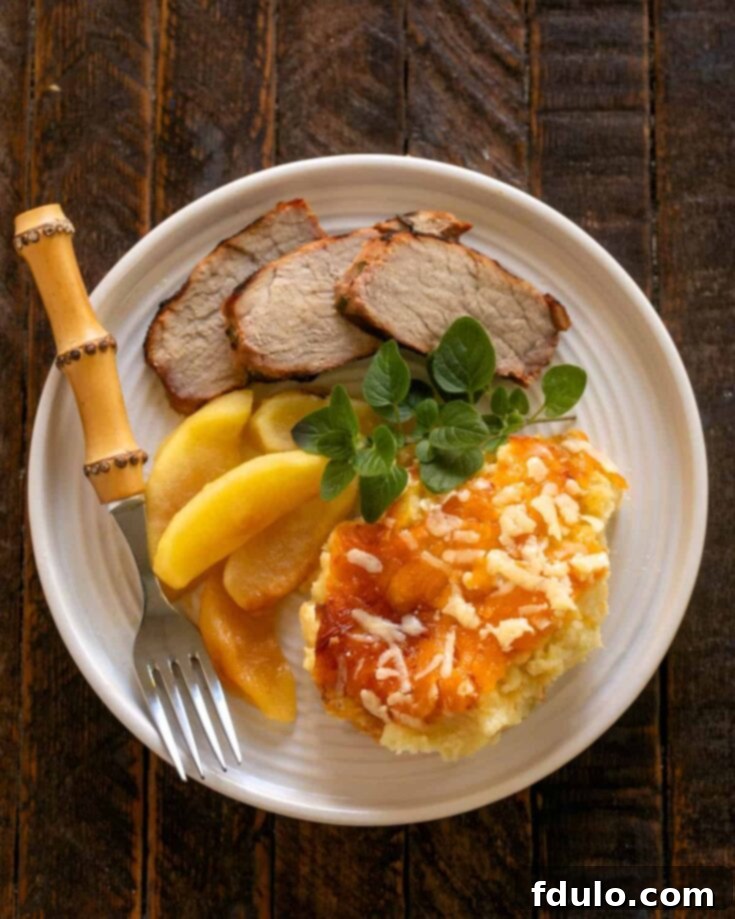 Overhead view of a golden brown, bubbly Twice Baked Potato Casserole, served on a white dinner plate with slices of pork and apples.