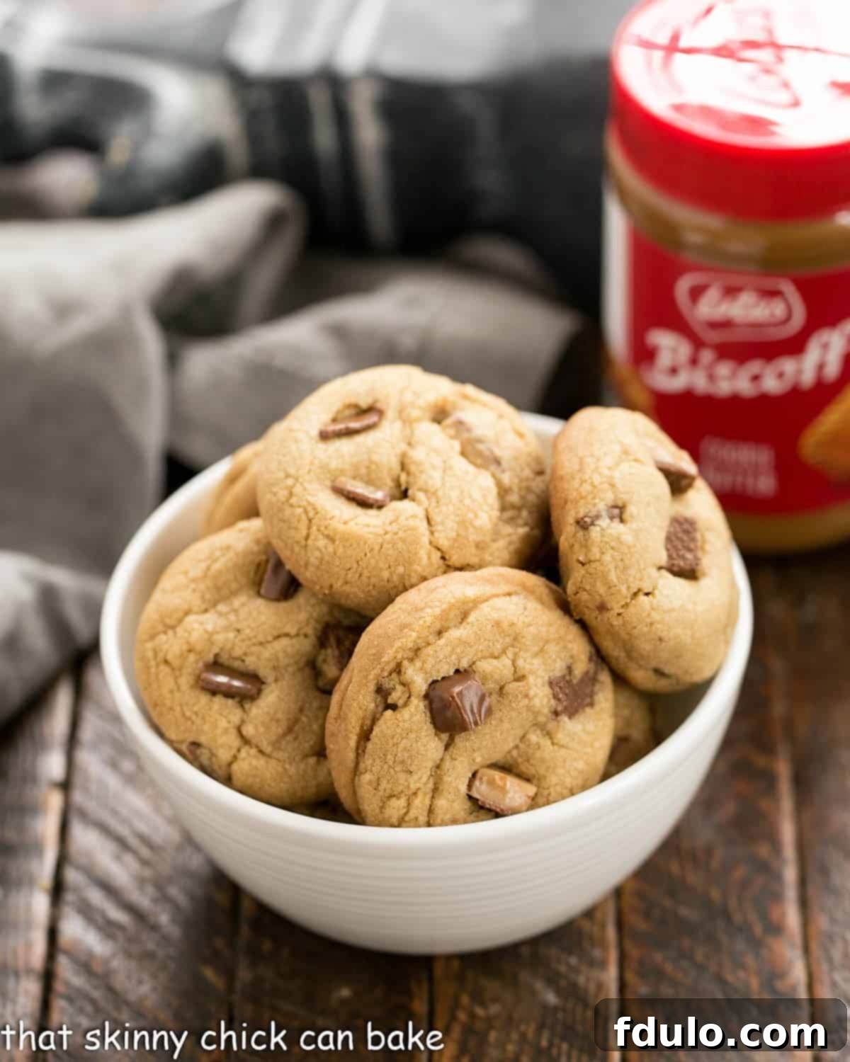 A beautifully arranged white bowl filled with golden-brown Toffee Biscoff Cookies, with a jar of creamy Biscoff cookie butter visible in the soft background.