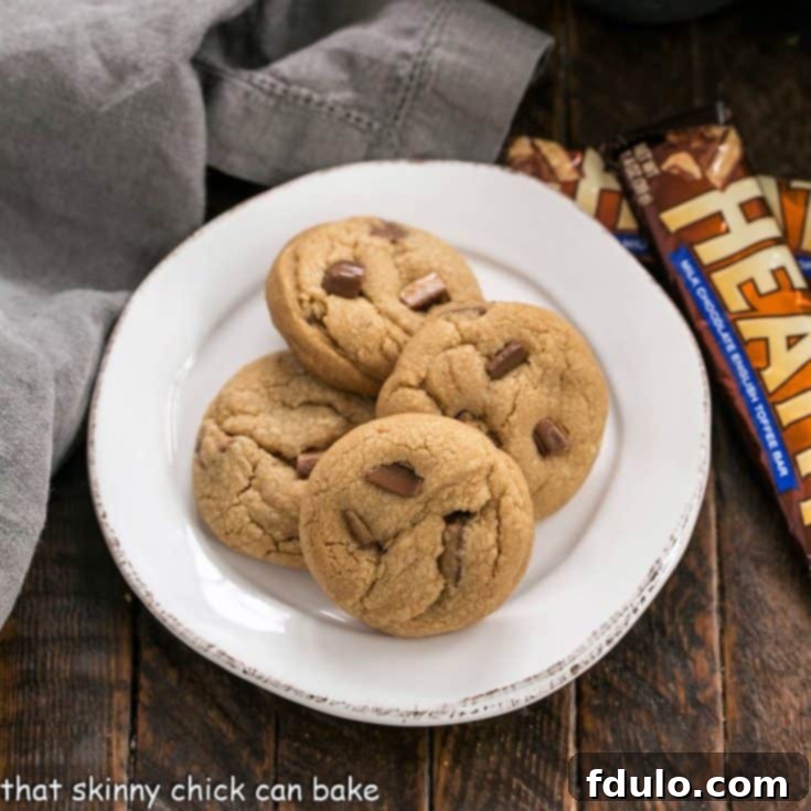 An overhead shot of golden-brown Biscoff Toffee Cookies neatly arranged on a white plate, with two Heath bars resting beside them, highlighting the key ingredient.