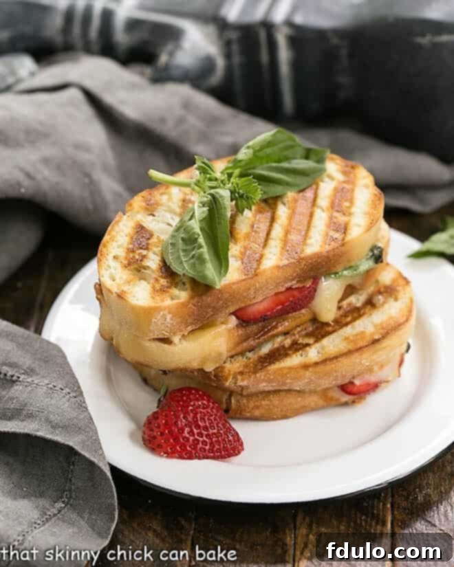 Gourmet Strawberry, Turkey, and Brie Grilled Cheese - an elevated, out-of-the-ordinary grilled cheese featuring gooey Brie, sweet strawberries, a hint of spicy jelly, and savory deli turkey! Half of a Strawberry, Turkey, and Brie Grilled Cheese Sandwich stacked on a round white plate, garnished with fresh basil and strawberry slices, showcasing the melted Brie and vibrant fillings.