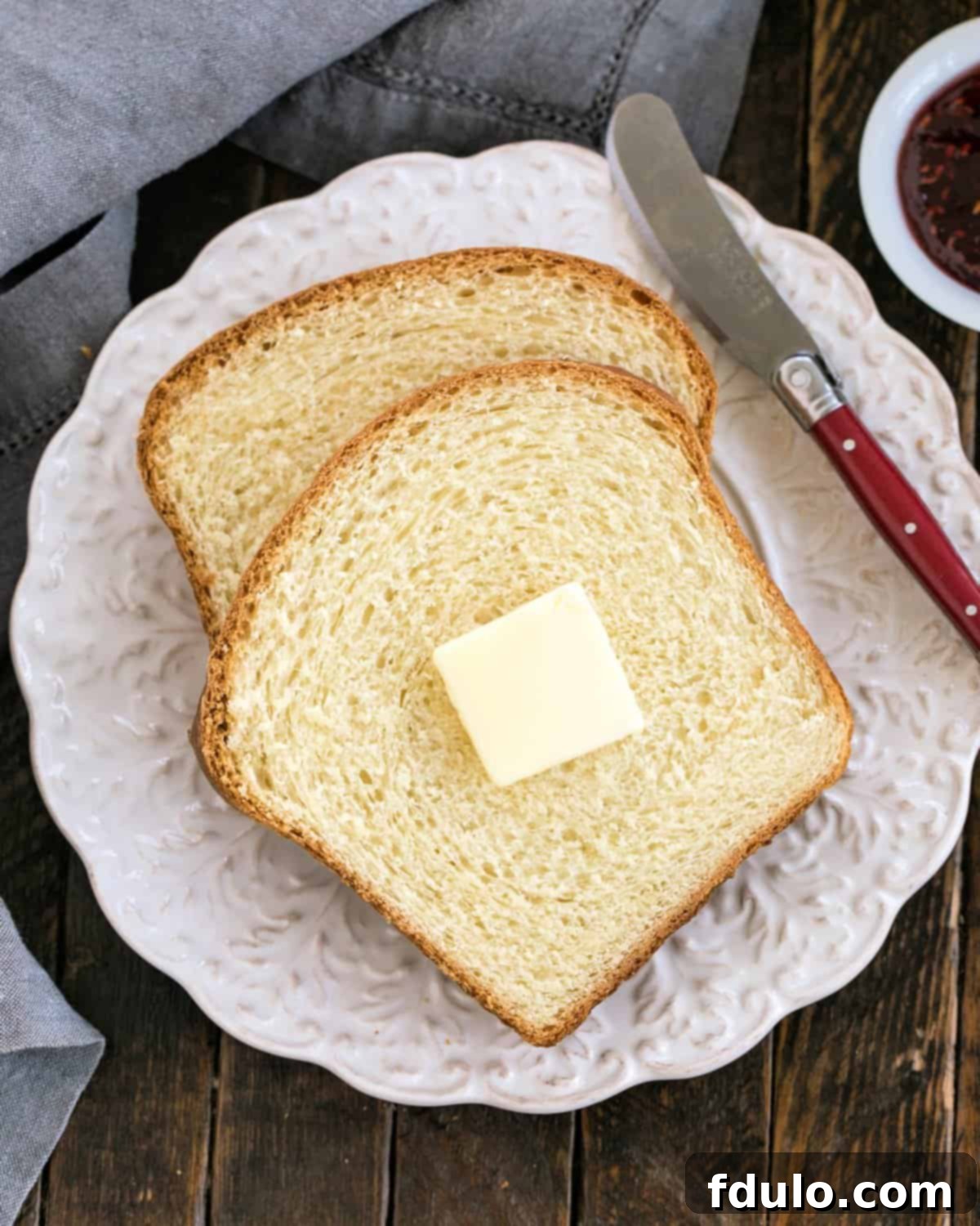 Two golden-brown slices of homemade potato bread with a pat of melting butter and a red-handled knife resting on a rustic white decorative plate.