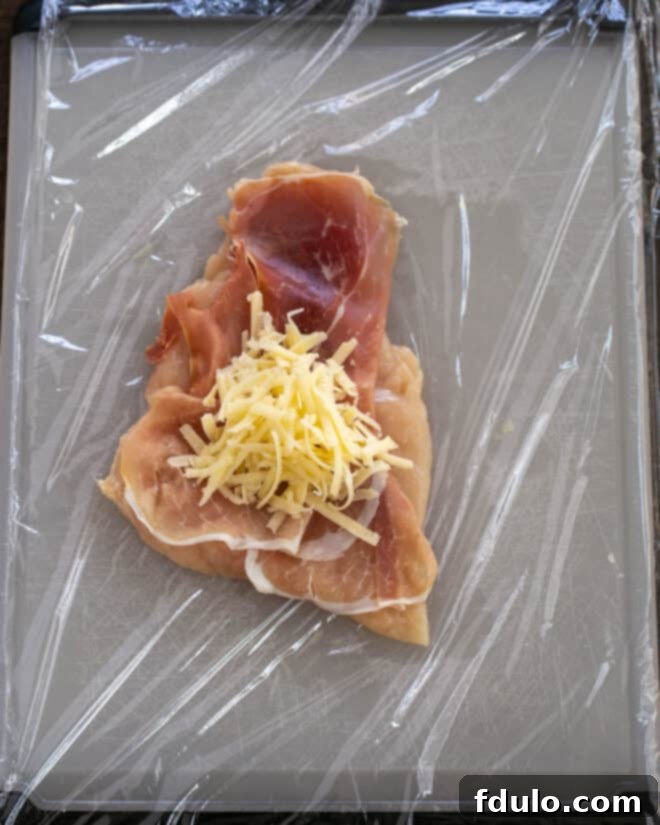 Step 2: A thin chicken cutlet laid flat on plastic wrap, topped with a slice of savory Prosciutto and a generous sprinkle of shredded Gruyere cheese, preparing for rolling.