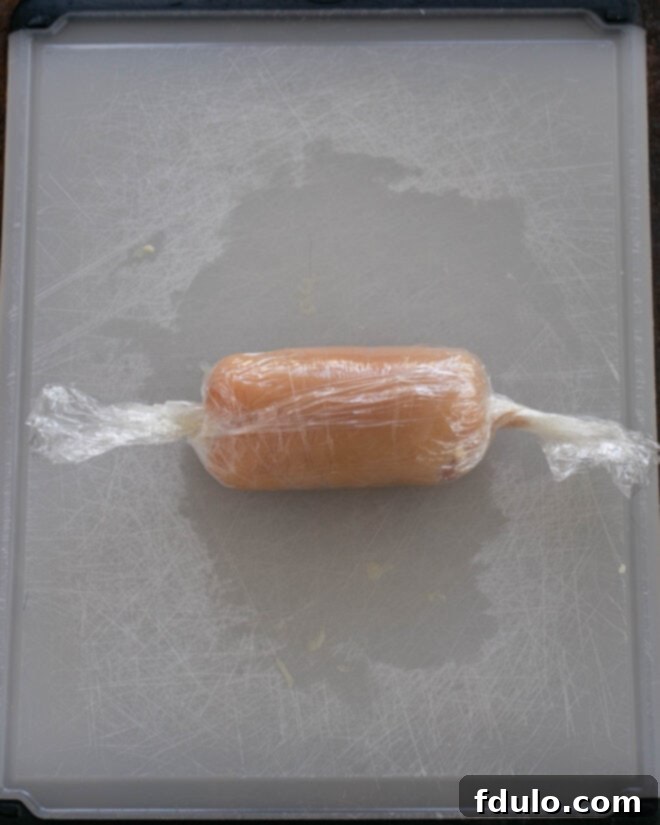 Step 3: A chicken cutlet, filled with prosciutto and cheese, being tightly rolled using plastic wrap to secure the ingredients and create a compact roll.