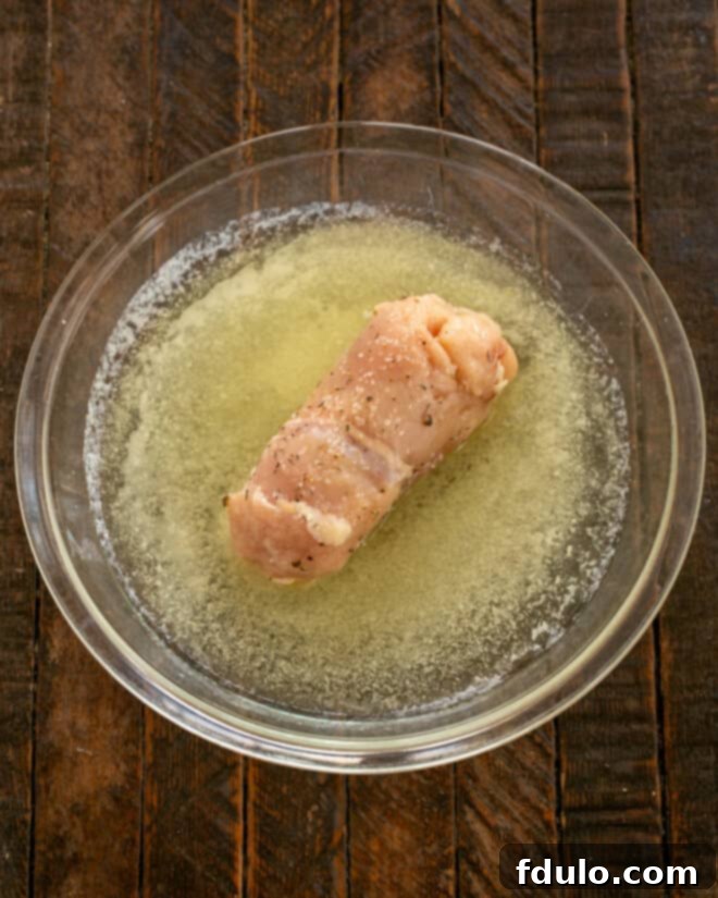 Step 5: A chilled, unwrapped chicken cordon bleu roll being dipped into a shallow dish of melted butter, ensuring an even coating before the breading step.