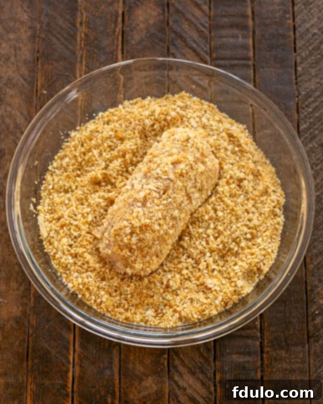 Step 6: A butter-coated chicken cordon bleu roll being generously rolled in a mixture of crushed seasoned croutons and grated Parmesan cheese, creating a thick, crispy breading.