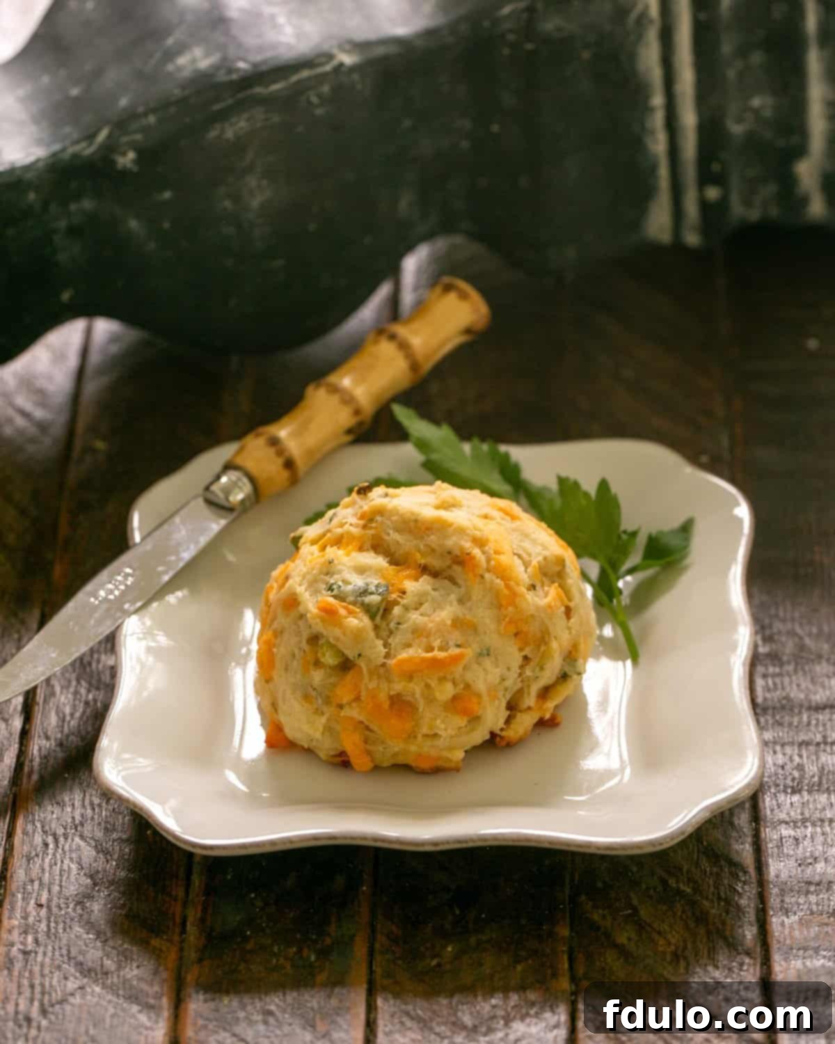 A golden-brown cheddar biscuit on a white plate, garnished with a sprig of fresh parsley, next to a butter knife, ready to be enjoyed.