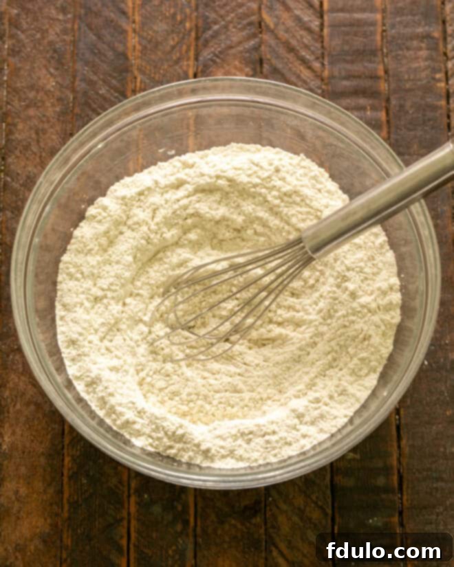 Dry ingredients for biscuits, including flour, baking powder, salt, sugar, and garlic powder, being whisked together in a large mixing bowl.