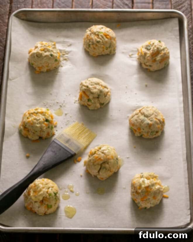 Scoops of cheddar biscuit dough, perfectly portioned, arranged on a parchment-lined baking sheet, ready for brushing with melted butter.