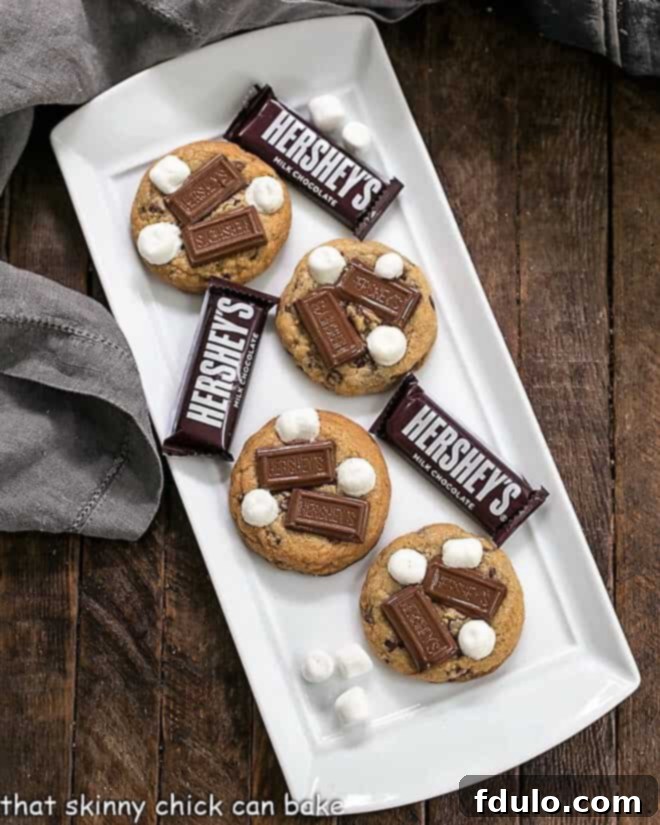 S'mookies AKA Gooey S'mores Cookies - All the amazing flavors of s'mores in gooey, chewy cookies S'mookies AKA Gooey S'mores cookies on a white ceramic tray, showing their golden edges and melted toppings.