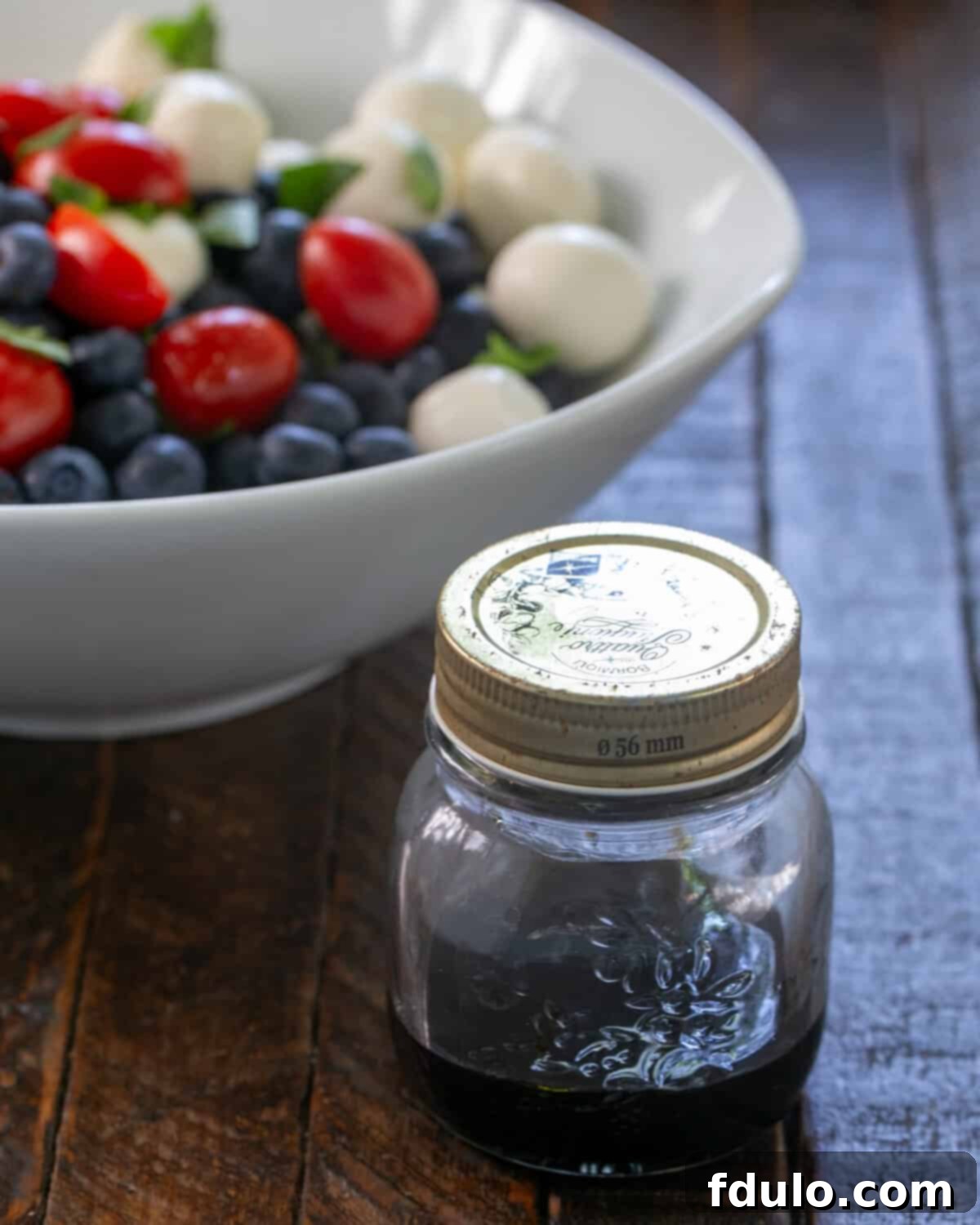 Blueberry Bliss Caprese 2 A vibrant Caprese Salad with Blueberries in a pristine white bowl, glistening with balsamic vinegar. Fresh basil leaves and small mozzarella balls are visible, promising a refreshing taste.