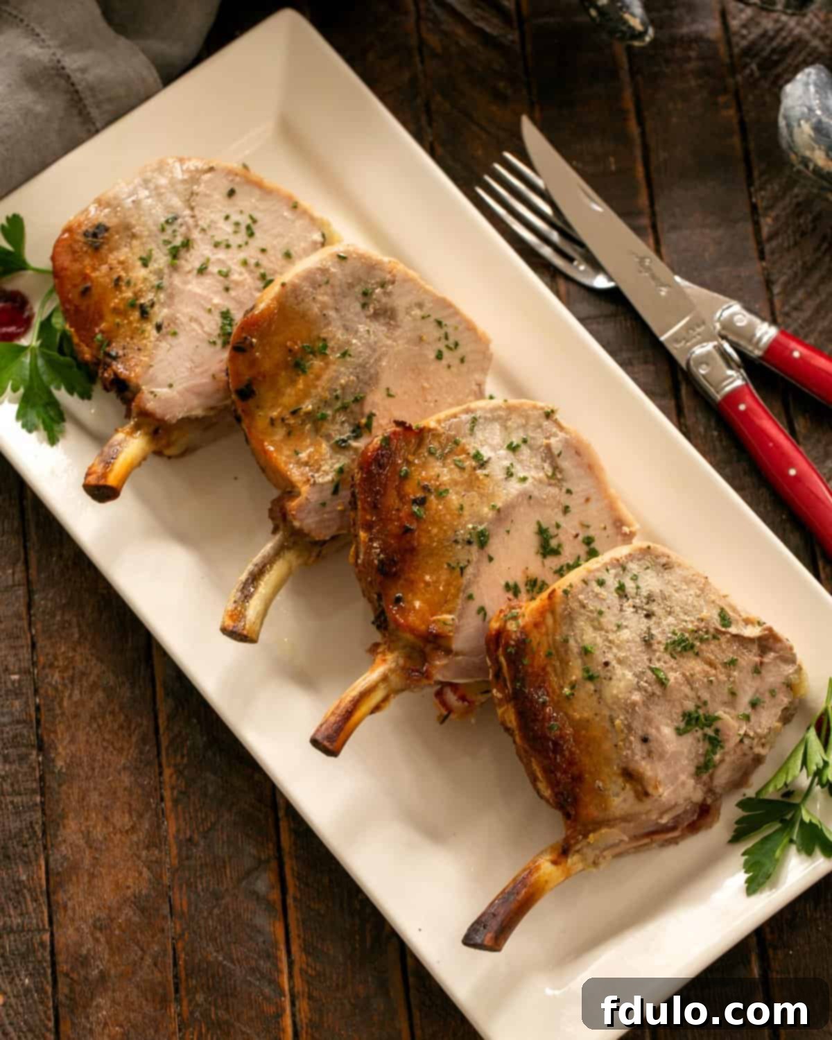 Beautifully sliced Crown Roast of Pork, garnished with fresh parsley, presented on an elegant white ceramic platter.