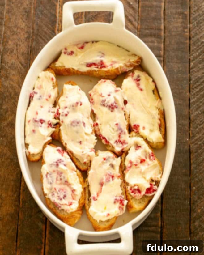 Flaky Croissant French Toast with Fresh Raspberries 6 The vibrant cream cheese and raspberry mixture being spread evenly and generously over the cut surfaces of the croissant bottoms already placed in the baking dish, creating a layer of filling.