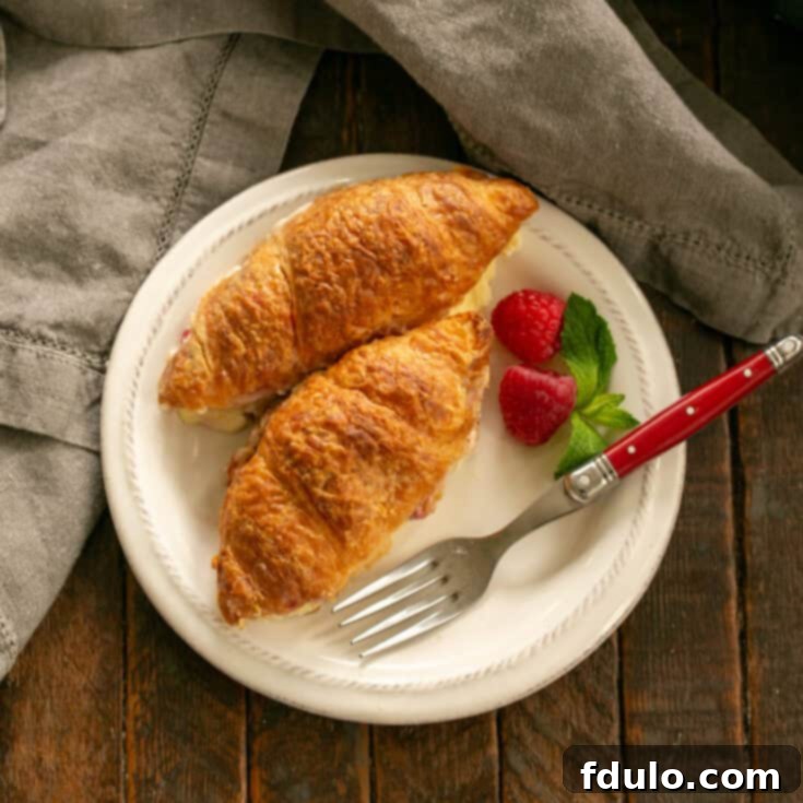 Flaky Croissant French Toast with Fresh Raspberries 10 Overhead view of a slice of Croissant French Toast on a white plate with fresh raspberry garnish and a dusting of powdered sugar, showing the creamy filling and flaky texture.