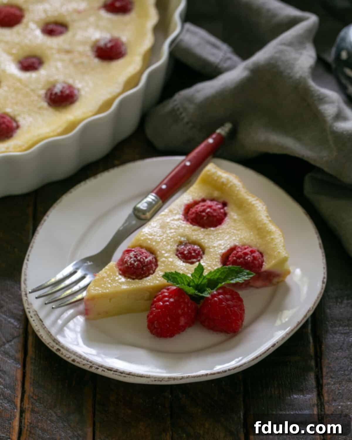 Slice of a raspberry clafoutis with a red handled fork on a white dessert plate with the serving dish in the background.